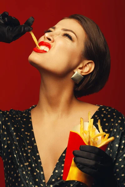 不健康的饮食 垃圾食品痴迷的概念 时尚豪华的年轻女子的肖像 在她的手吃油炸土豆 摆在红色背景 关门了工作室拍摄 — 图库照片