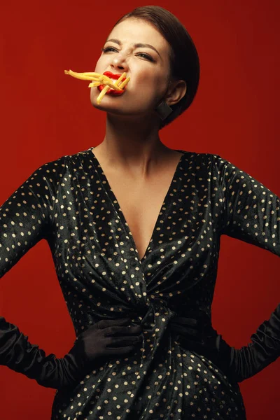 不健康的饮食 垃圾食品痴迷的概念 时尚豪华的年轻女子手持的肖像 吃油炸土豆 在嘴里 摆在红色背景 工作室拍摄 — 图库照片