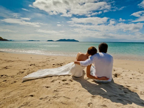 Gelin ve damat düğün elbiseli okyanusun üzerinde oturan ve uzaktan izliyor. Arkadan görüntülemek.