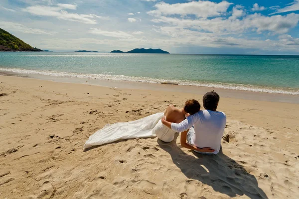 Gelin ve damat düğün elbiseli okyanusun üzerinde oturan ve uzaktan izliyor. Arkadan görüntülemek.