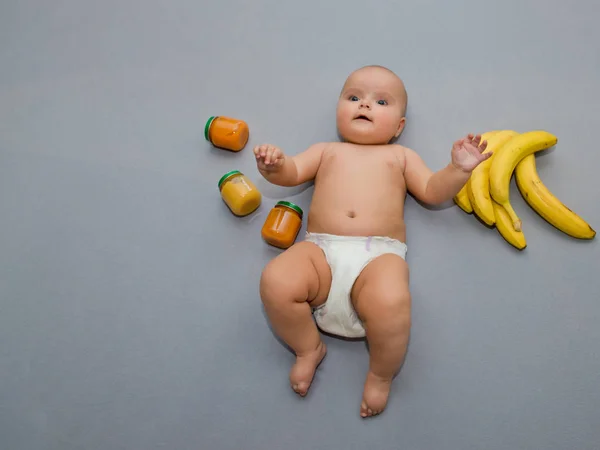 Überraschtes Baby Auf Grauem Hintergrund Mit Bananen Und Gläschen Babynahrung — Stockfoto