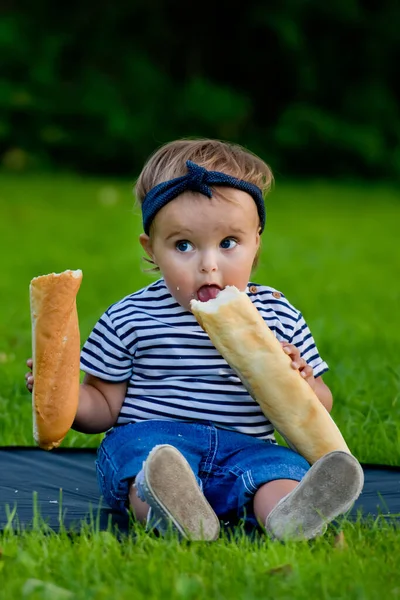 一个漂亮的小女孩坐在花园里的草坪上 手里拿着一个新鲜的面包 — 图库照片