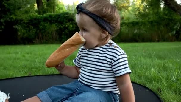 Маленькая хорошенькая девочка сидит на лужайке в саду и держит свежий багет. — стоковое видео