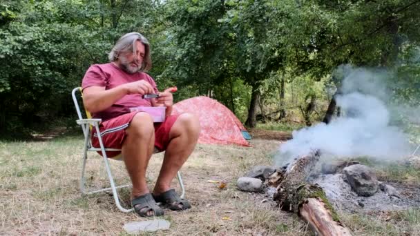 Dorosły mężczyzna z brodą siedzi przy ogniu i robi papierosa z tytoniem.. — Wideo stockowe