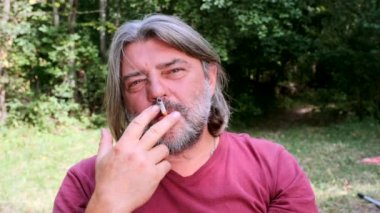 Doğanın arka planında sigara içen yetişkin sakallı bir adamın portresi. Yavaş çekim.