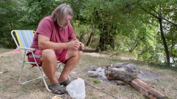 ひげを生やした漁師は、観光キャンプでキャンプファイヤーの近くに座って釣りの餌を準備する. — ストック動画