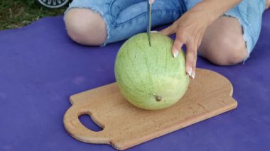 Kadın, bahçedeki mavi bir halıda otururken bir karpuzu tahtaya ikiye böler. Yakın plan..