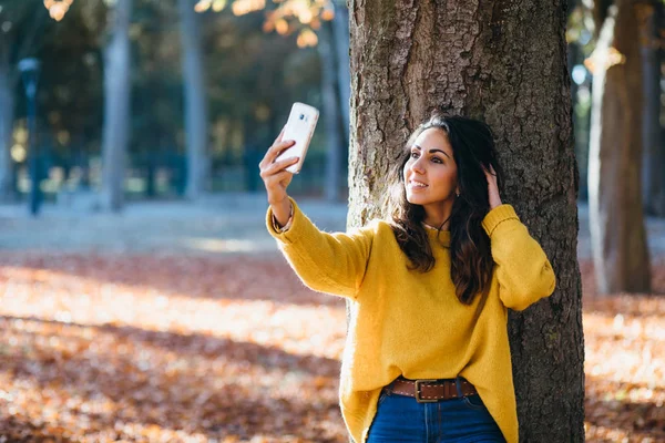Casual beautiful young woman posing for smartphone selfie photo and touching her hair at city park in autumn.