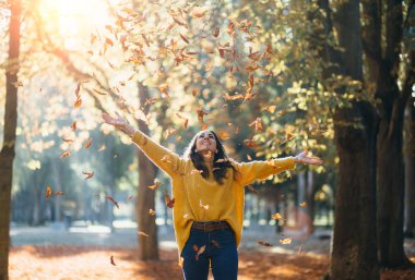Casual joyful woman having fun throwing leaves in autumn at city park. clipart