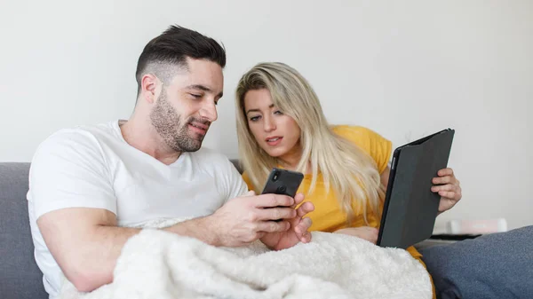 Νεαρό περιστασιακό ζευγάρι χρησιμοποιώντας κινητά τηλέφωνα και tablet στο σπίτι — Φωτογραφία Αρχείου