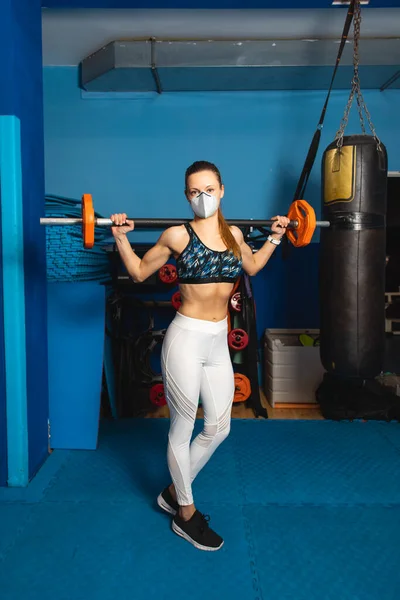 Young fit woman with n95 face mask  exercising at the gym with weights. Fitness strength workout under coronavirus health crisis.