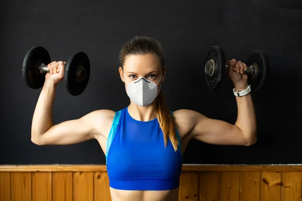年轻健康的女人戴着N96口罩 用哑铃按肩膀 在体育馆里锻炼 验尸官健康危机下的健身力量锻炼 — 图库照片