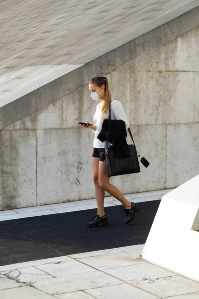 Businesswoman wearing n95 face mask for protecting against coronavirus. Young stylish professional woman walking on the street and texting on smartphone.