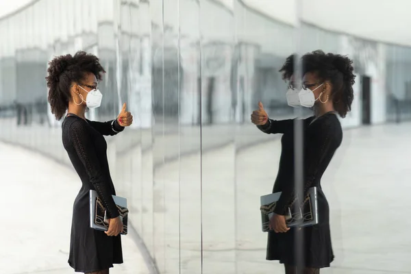 Successful stylish businesswoman with n95 face mask doing thumbs up gesture to herserlf in glass reflection. Professional black woman giving luck to herself under coronavirus Covid.19 health crisis.