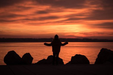 Okyanusun üzerine düşünceler ile canlı turuncu deniz günbatımı bakan kayaların üzerinde kontrast duran genç çocuk.