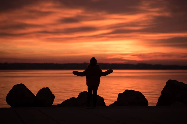 年轻的孩子站在岩石的轮廓俯瞰一个生动的橙色海洋日落与海洋的反射 — 图库照片