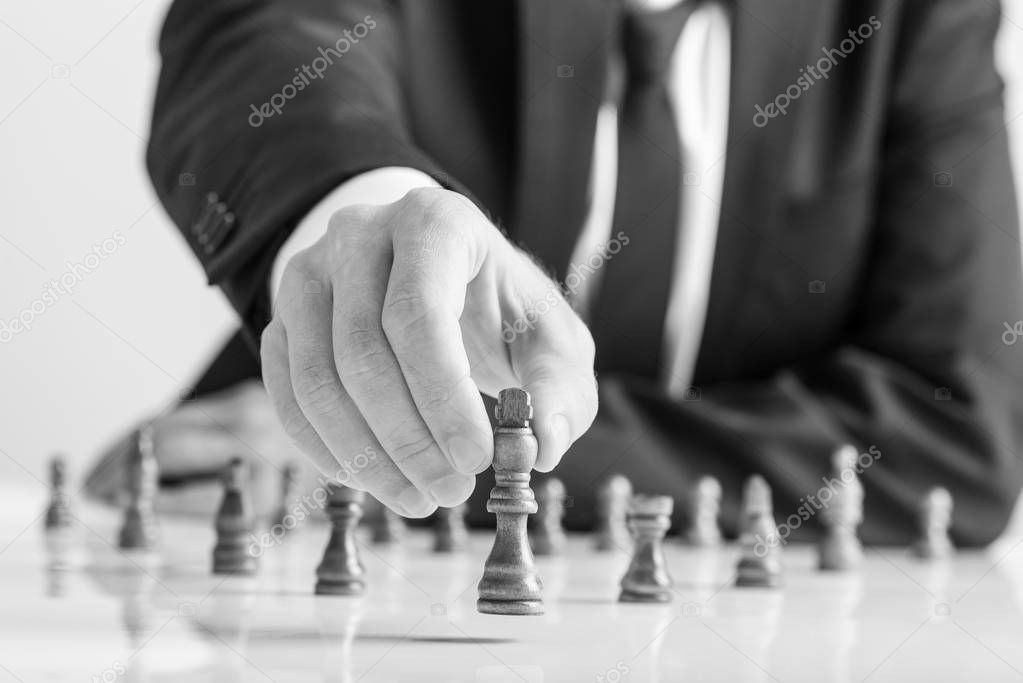 Greyscale image of businessman wearing business suit moving dark King chess piece at white table. Conceptual of progress and success.