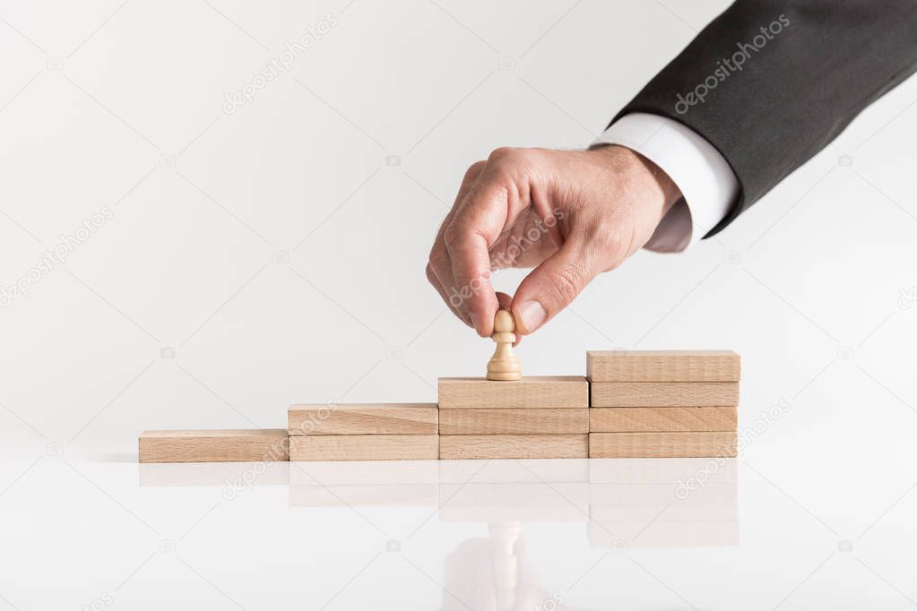 Conceptual image of career management with a businessman placing pawn chess piece on a staircase of wooden bricks over white background.
