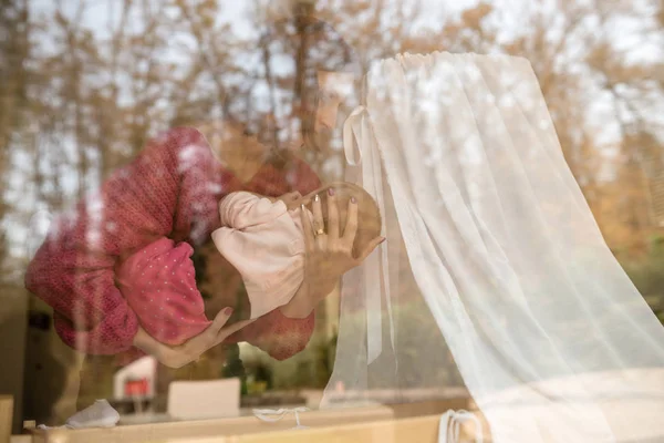 妇女抱着一个婴儿在家中 旁边的婴儿床通过一个大的平板玻璃窗口 与树木的反射 — 图库照片