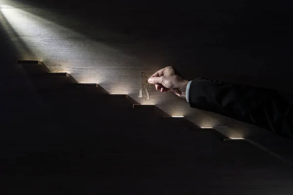 Businessman climbing the steps to success with paper silhouette cutouts of a man and a human hand helping him climb towards the light.