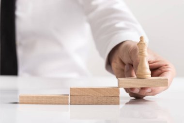 Businessman holding a queen chess piece on a wooden block positioned as the top in a row of steps in a concept of teamwork, success, and achievement. clipart