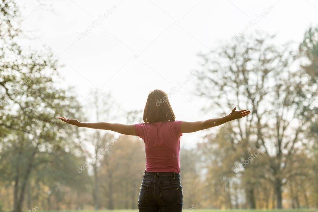 View from behind of a young woman standing in park with her arms spread widely looking towards the trees.