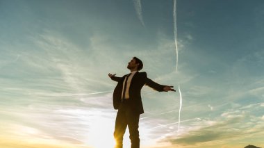 Başarılı iş adamı ayakta göklere bakarak uzanmış kollarıyla güneşin parlak parlaklık karşı kontrast gün batımında sevindirici.