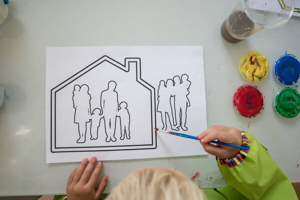 Top view of toddler boy or girl colouring a house with family silhouette printed on white paper.