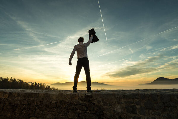 Businessman holding aloft his jacket at sunset as he stands on a wall overlooking a misty mountain scene silhouetted against the bright colorful sky with contrails.