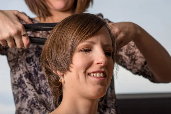 一个年轻的微笑的女人坐在理发师的肖像 而她正在矫直她的棕色头发 — 图库照片