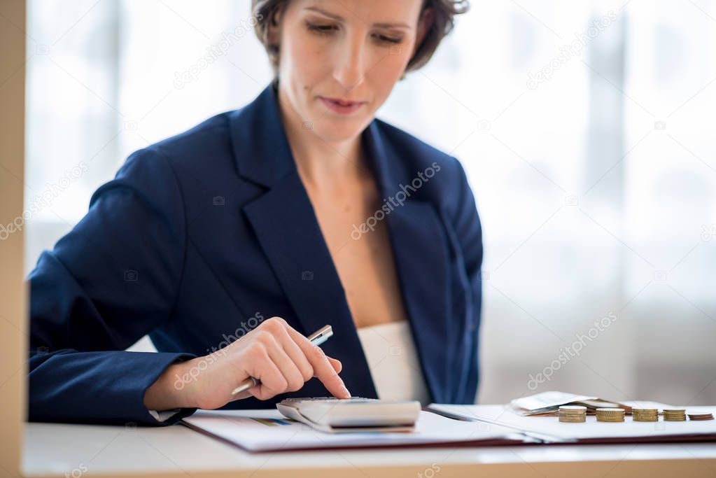 Front view of pretty young business woman sitting behind her desk calculating income with stacks of coins placed on paperwork she is reviewing.