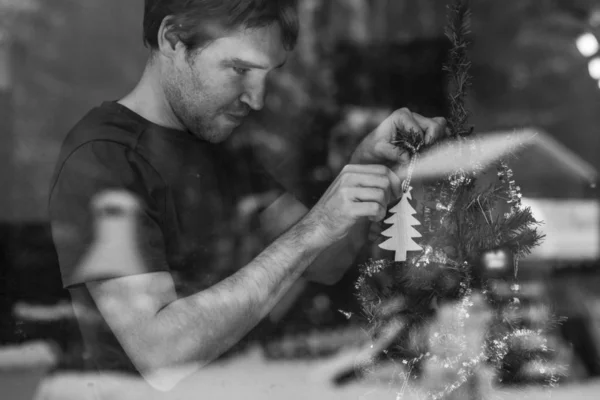 Black and white image of a young man hanging holiday decoration on a Christmas tree. View through a window with snowy nature reflecting in the glass.