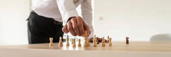 Business leadership concept - businessman standing at his office desk placing chess figures on table with the king in front.