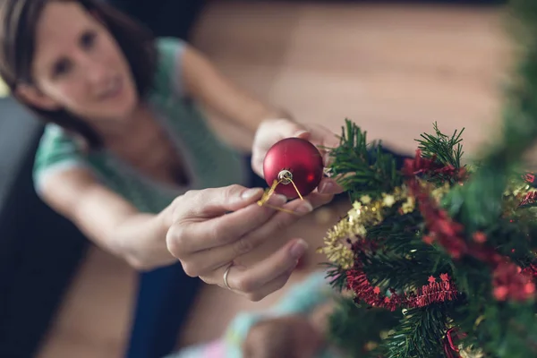 Retro vintage image of a young woman sitting on the floor placing red Christmas bauble on small holiday tree.