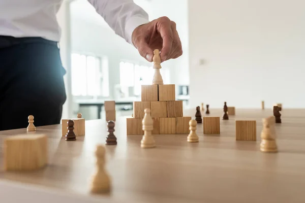 Business hierarchy concept with businessman placing chess figure of king on top of wooden stacked wooden blocks and other figures spread on office desk.