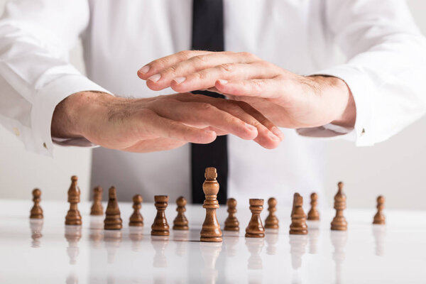 Бизнесмен, держащий в руках защитные руки над шахматными фигурами, размещенными на офисном столе с королем в ведущей позиции
.