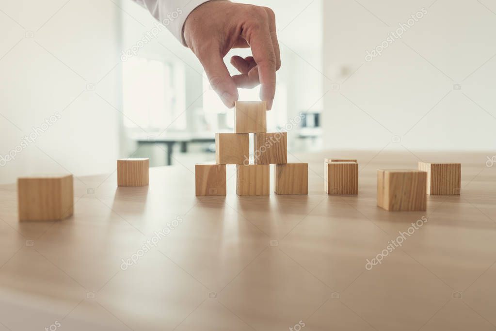 Businessman placing wooden cubes in a pyramid shape on office desk in his workplace.