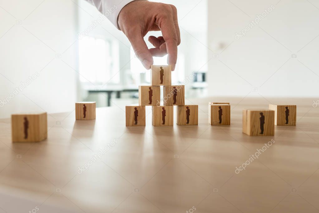 Businessman arranging wooden cubes with people silhouette in a pyramid shape with others scattered on office desk. Conceptual of business organization, human resources and teamwork.