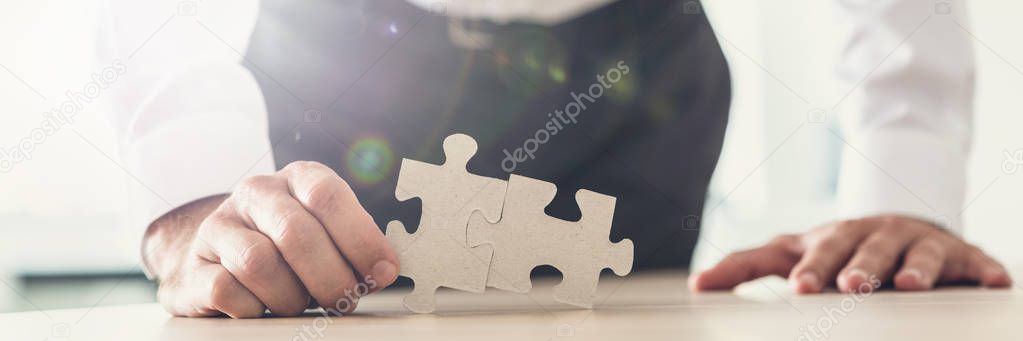 Wide view image of a businessman holding two puzzle pieces matched together as he leans on office desk. With lens flare.