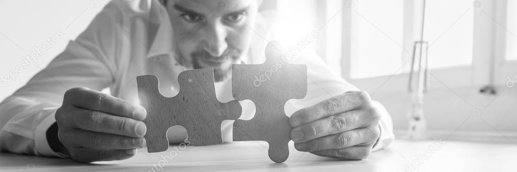 Black and white wide view image of businessman leaning on his office desk matching two wooden puzzle pieces with lens flare.