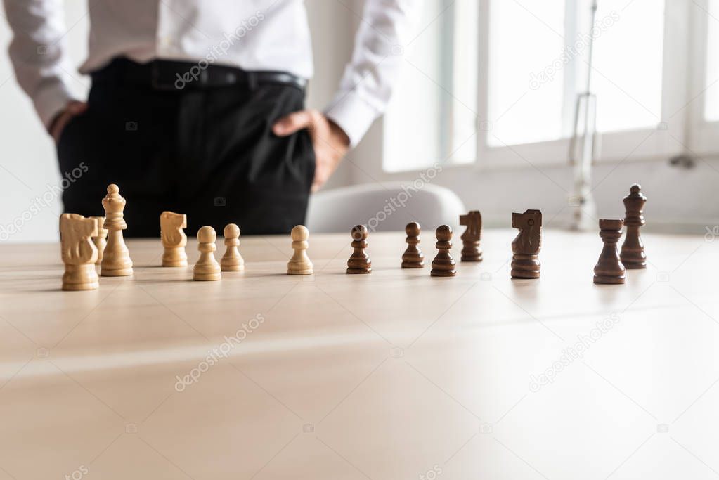 Businessman standing at his office desk looking at black and white chess pieces arranged on the wooden desk. With copy space on the lower part of the image.
