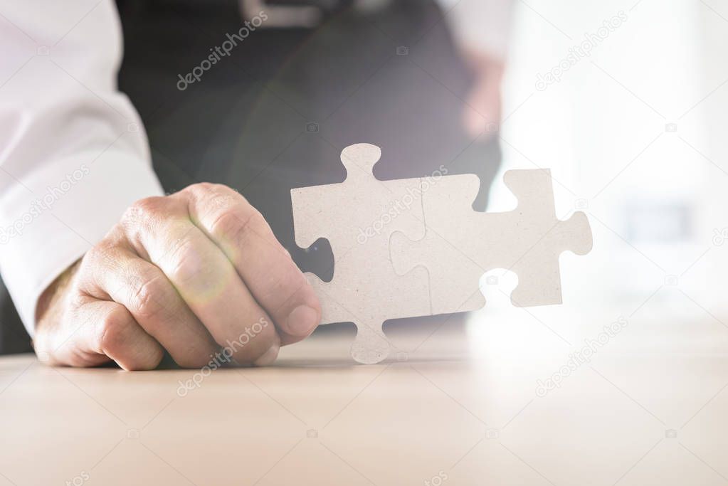 Businessman holding two joined matching puzzle pieces leaning on his office desk. With bright background and lens flare.