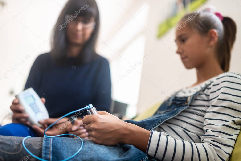 Teenage girl holding zapper electrodes as she attends a bioresonance therapy with her female therapist.
