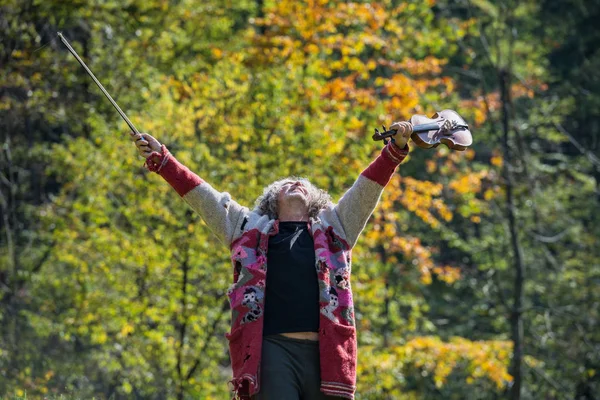 バック グラウンドで彼のヴァイオリンと弓の木が外の空気を高を持ち上げる人生を祝って破れた古いセーターで年配のホームレス男性 — ストック写真