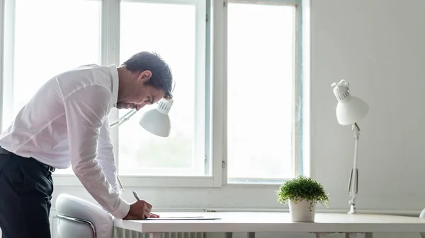 ランプとそれの緑の植物契約または白のオフィスの机の上のドキュメントに署名するに傾いて執務室の机に立っているビジネスマン — ストック写真