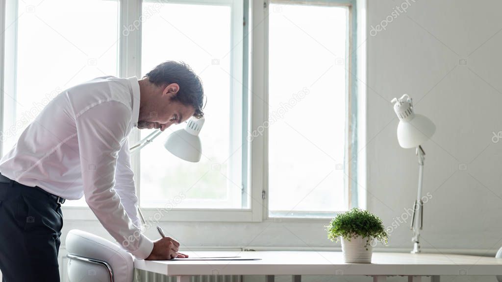 Businessman standing at his office desk leaning in to sign a contract or document on white office desk with a lamp and green plant on it.