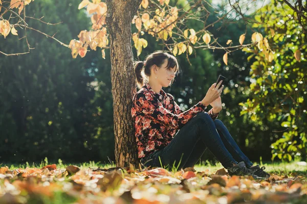 Junge Brünette mit ihrem Handy unter einem Baum sitzend — Stockfoto