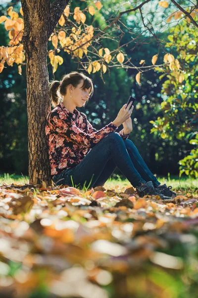 Žena, která sedí pod stromem a prohlížeje si mobil — Stock fotografie