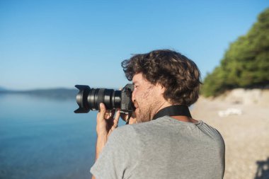 Genç erkek fotoğrafçı sabahın erken saatlerinde profesyonel DSR kamerasıyla güzel deniz ve doğanın fotoğraflarını çekiyor..