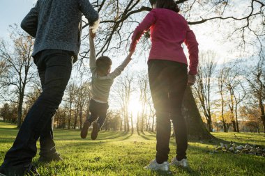 Genç ebeveynlerin, oğullarının ellerini tutarak parkta güneşin altında yürürken onu neşeli bir şekilde kaldırışlarına bakın..